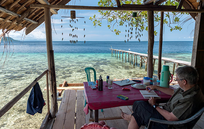 A man sits with a notebook at a table on a platform overlooking a reef in Raja Ampat